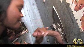 An ebony woman in the USA performs an interracial gloryhole blowjob, sucking a big black cock through the hole